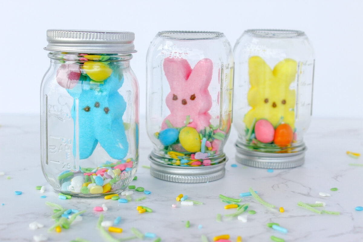 Three mason jars filled with colorful marshmallow bunnies, eggs, and sprinkles are displayed on a marble surface.