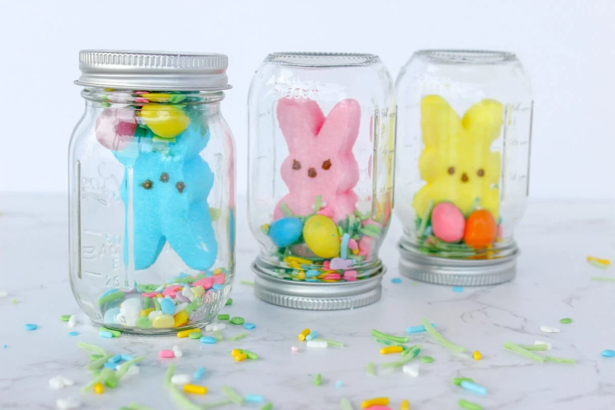 Three mason jars filled with colorful marshmallow bunnies, eggs, and sprinkles are displayed on a marble surface.