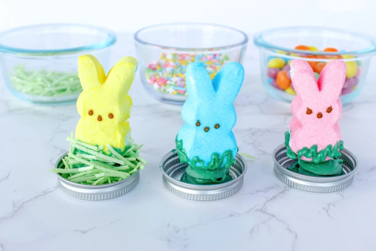 Three marshmallow bunny treats in yellow, blue, and pink rest on green coconut grass nestled in jar lids. Behind them, glass bowls with sprinkles and jelly beans evoke a whimsical Easter snow globe scene on the marble surface.