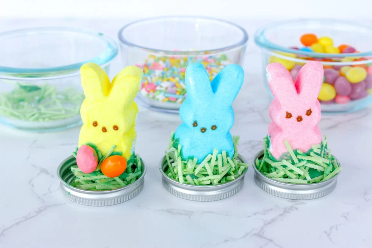 Three marshmallow bunnies in yellow, blue, and pink sit like Easter snow globes on jar lids with green shredded grass. They're surrounded by colorful candy on a white surface, with jars of sprinkles and jelly beans adding to the festive scene.