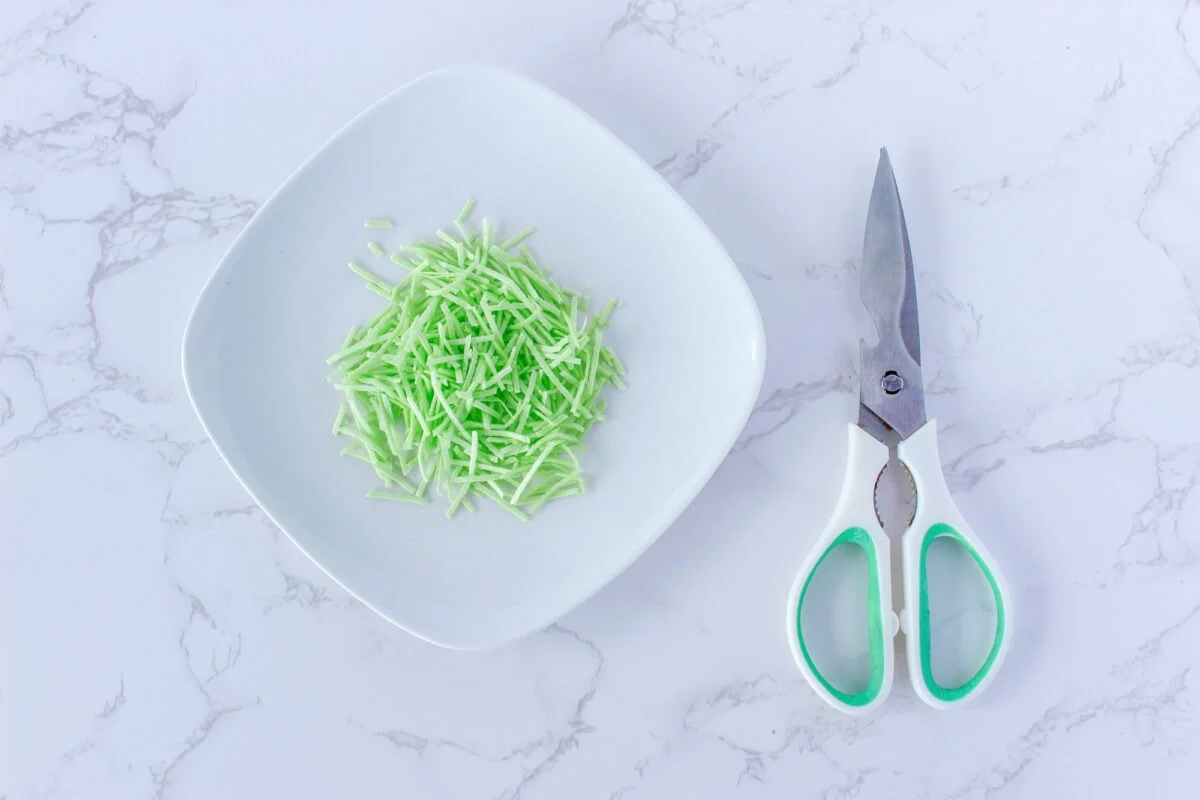 White square plate with shredded green paper next to green-handled scissors on a marble surface.