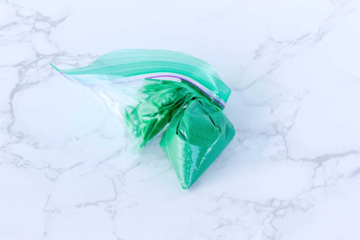 A clear plastic bag filled with green frosting, sealed and twisted at the top, placed on a marble surface.