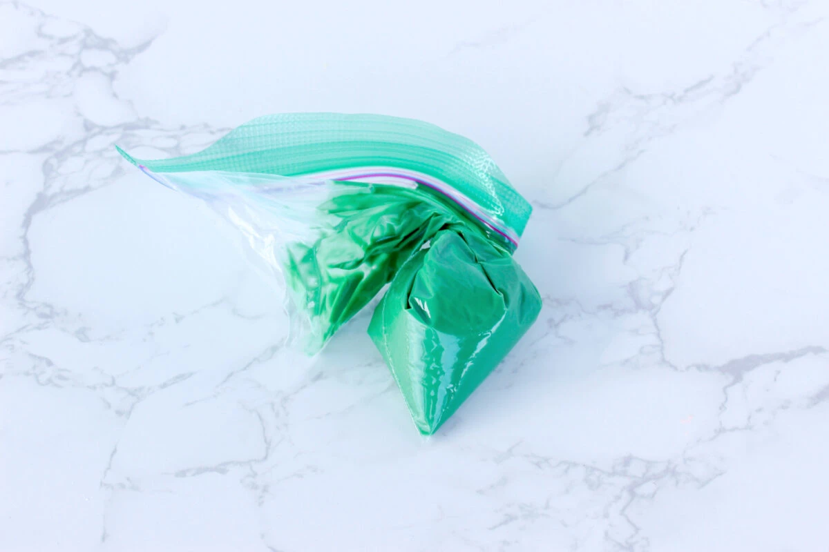 A clear plastic bag filled with green frosting, sealed and twisted at the top, placed on a marble surface.