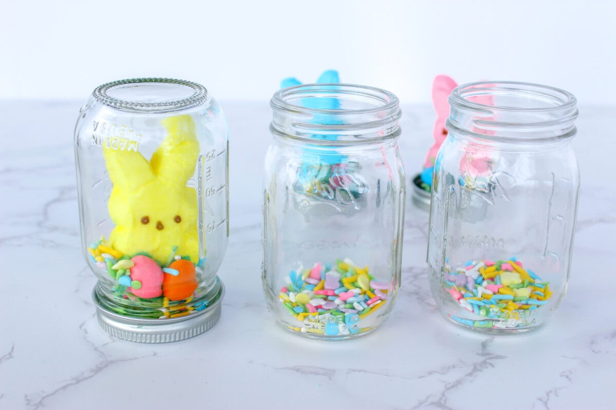 Three glass jars with colorful marshmallow bunnies and festive sprinkles inside, arranged on a white marble surface.