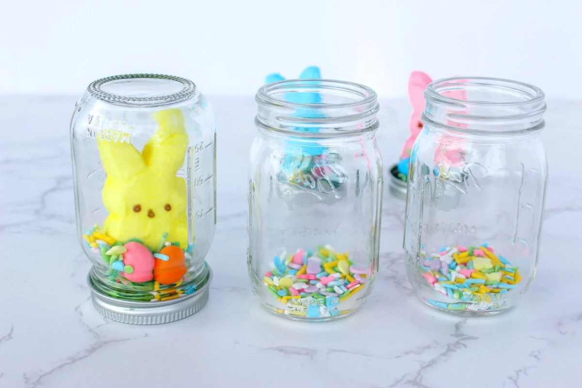 Three glass jars with colorful marshmallow bunnies and festive sprinkles inside, arranged on a white marble surface.