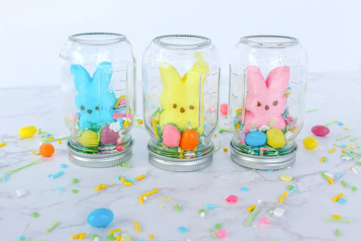 Three glass jars are inverted on lids, each containing a colored marshmallow bunny and assorted candies. The jars are surrounded by scattered candy on a marble surface.