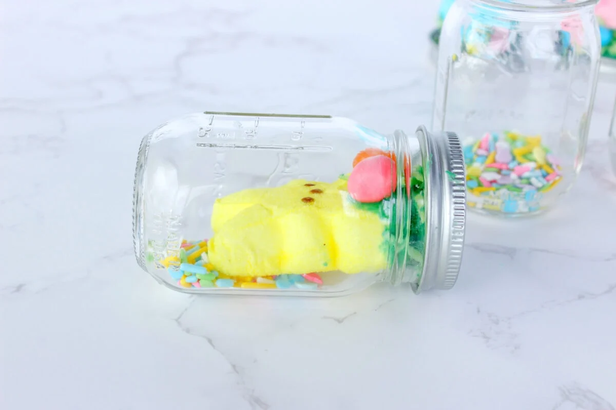 A glass jar on its side cradles a small yellow chick sponge figure, resembling one of those playful Easter snow globes, surrounded by colorful sprinkles on a marble surface.
