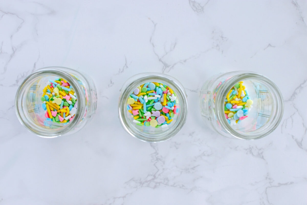 Three glass jars filled with colorful sprinkles on a white marble surface.