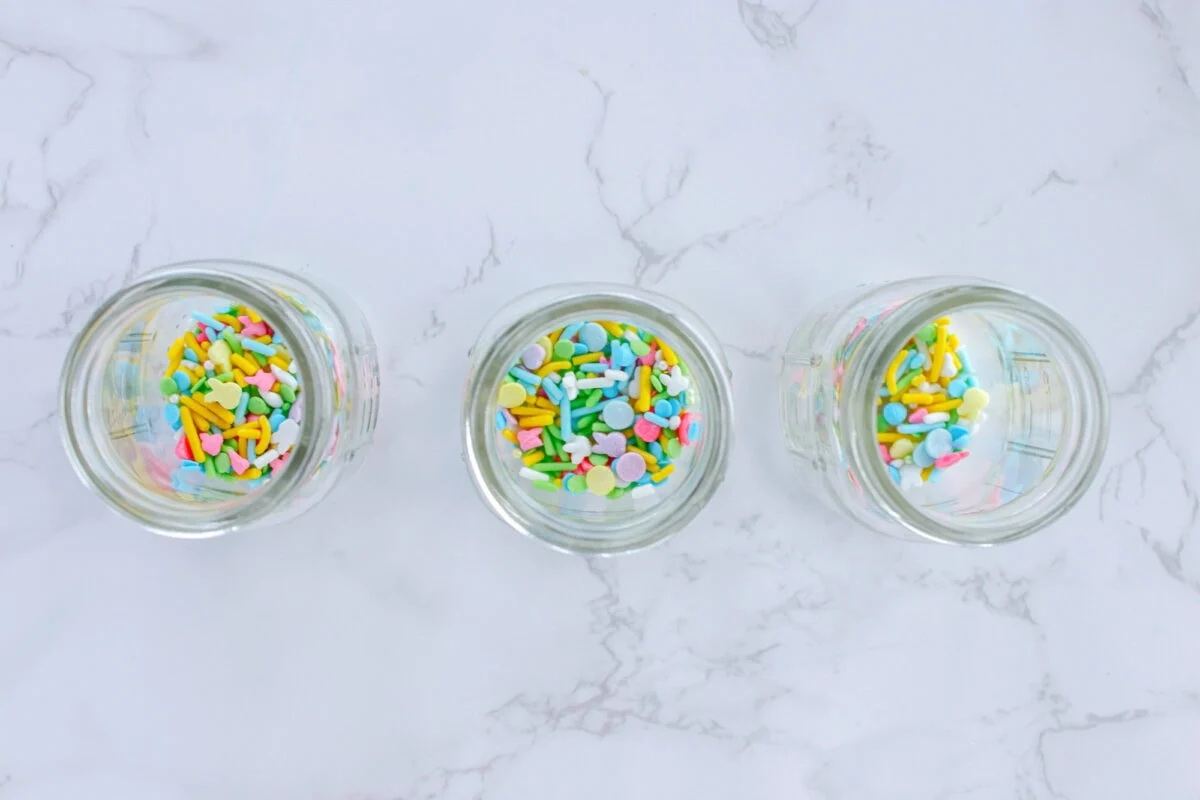 Three glass jars filled with colorful sprinkles on a white marble surface.