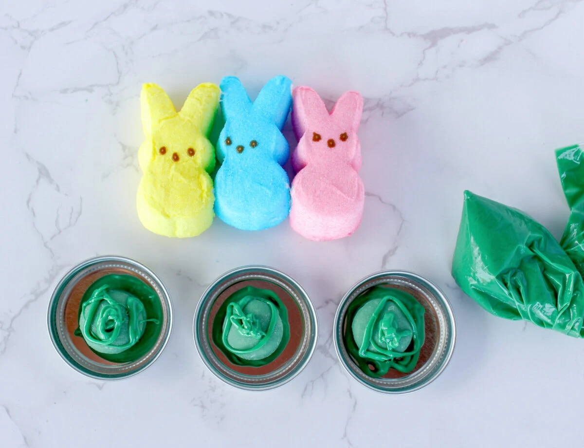 Three marshmallow bunnies in yellow, blue, and pink sit above three small jars with green icing against a marble background. A green icing bag is on the right.
