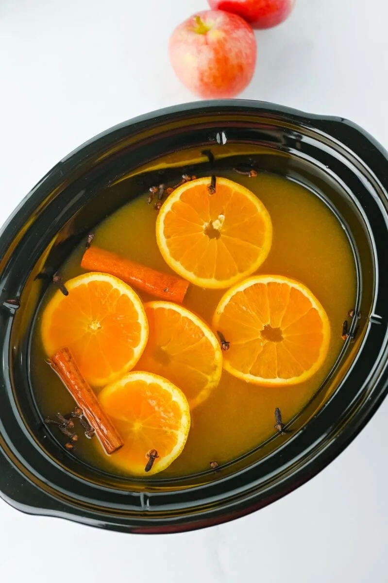 A slow cooker filled with orange slices, cinnamon sticks, and cloves in liquid, with apples in the background on a white surface.