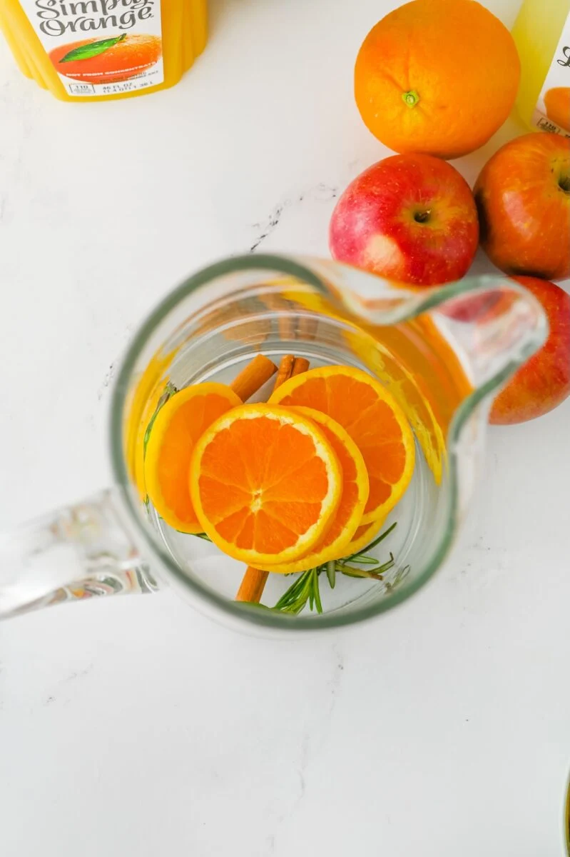 A glass pitcher with orange slices, cinnamon sticks, and rosemary, next to apples, an orange, and a container of orange juice on a white surface.