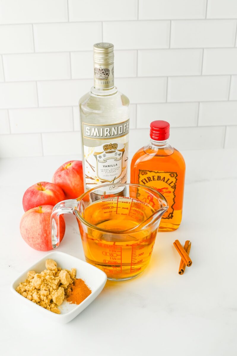 A bottle of vanilla vodka, a bottle of cinnamon whiskey, apples, a measuring cup with liquid, ground spices in a dish, and cinnamon sticks on a white countertop.