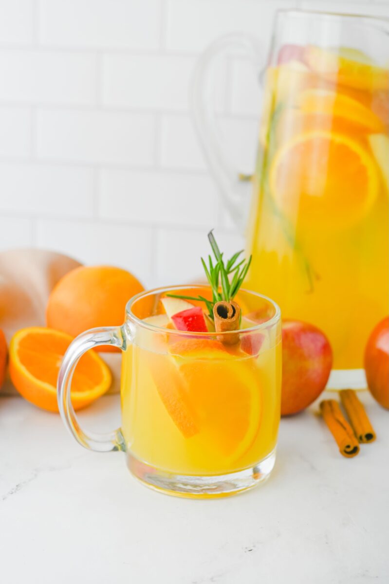 A glass mug of citrus and apple-infused yellow drink garnished with rosemary and a cinnamon stick, with oranges and apples in the background and a pitcher of the same drink.