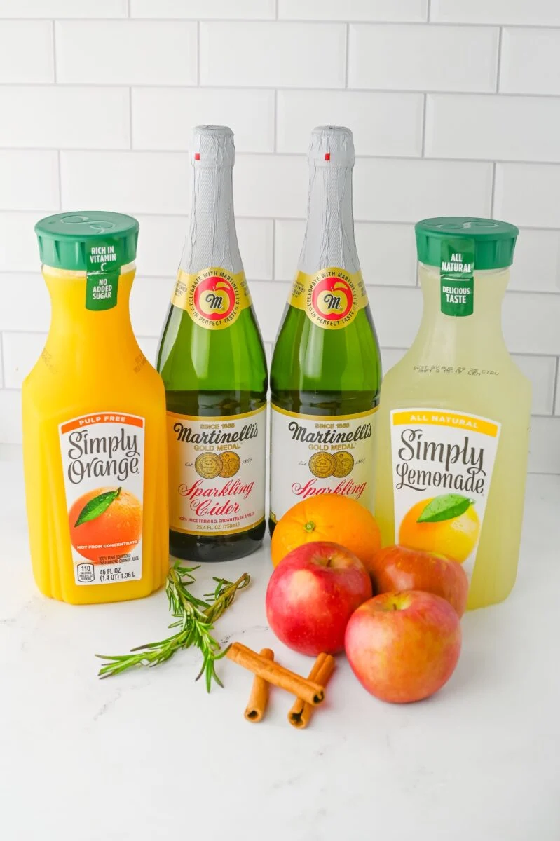 Bottles of Simply Orange and Simply Lemonade, two Martinelli’s Sparkling Cider bottles, three apples, cinnamon sticks, and rosemary on a white surface with a tiled background.