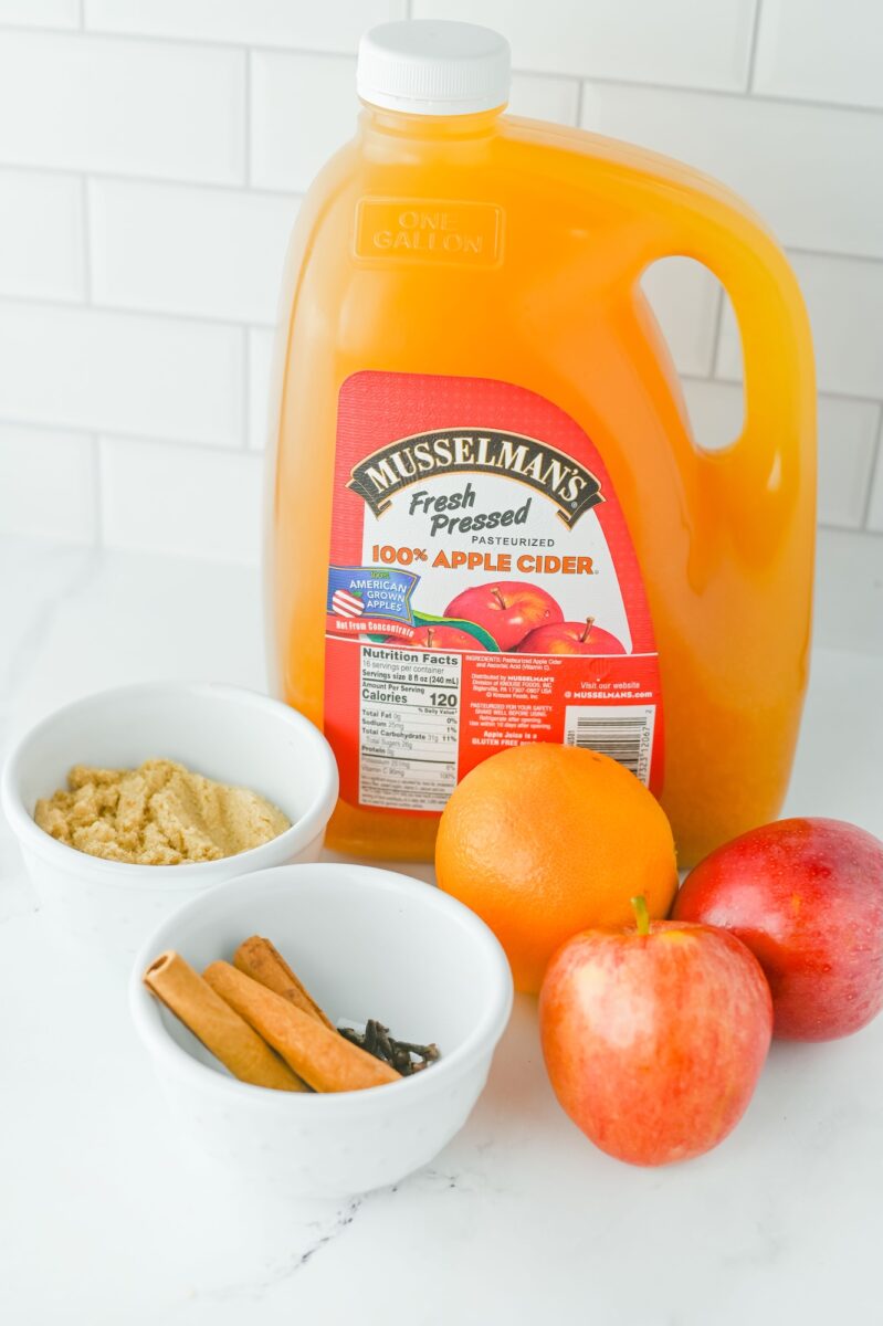 A gallon jug of Musselman’s apple cider next to bowls of brown sugar and spices, with an orange and two red apples on a white countertop.