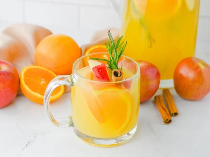 A clear glass mug filled with yellow citrus drink, garnished with orange slices, apple pieces, rosemary, and a cinnamon stick; apples, oranges, and cinnamon sticks in the background.