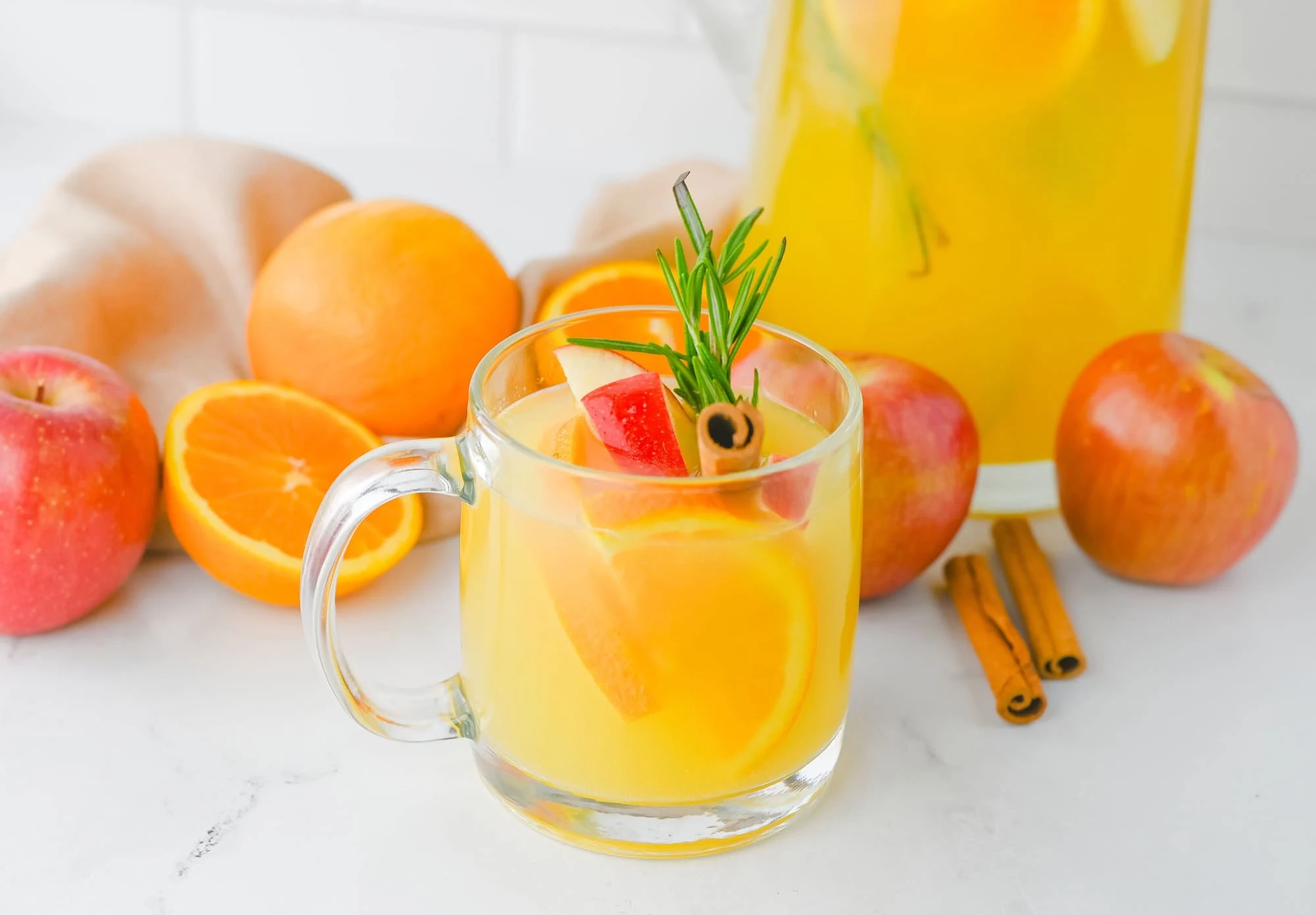 A clear glass mug filled with yellow citrus drink, garnished with orange slices, apple pieces, rosemary, and a cinnamon stick; apples, oranges, and cinnamon sticks in the background.