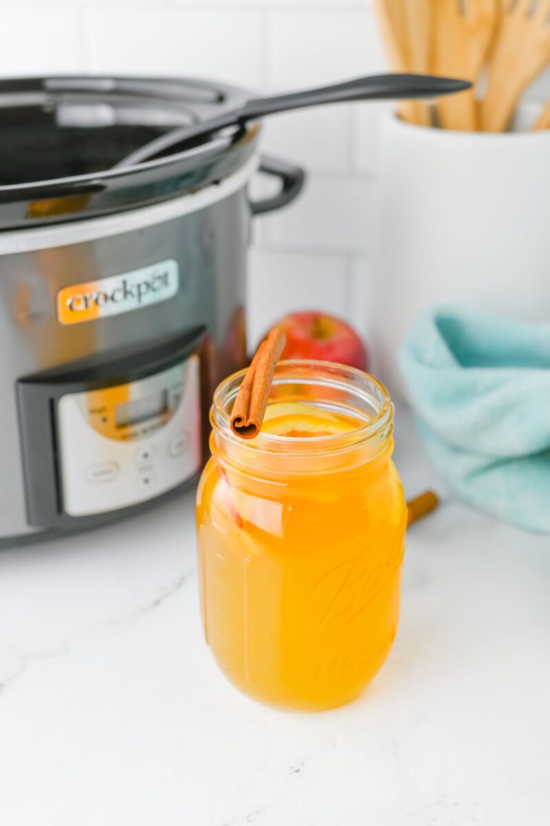 A mason jar filled with orange-yellow liquid and a cinnamon stick is placed in front of a slow cooker on a white kitchen counter.