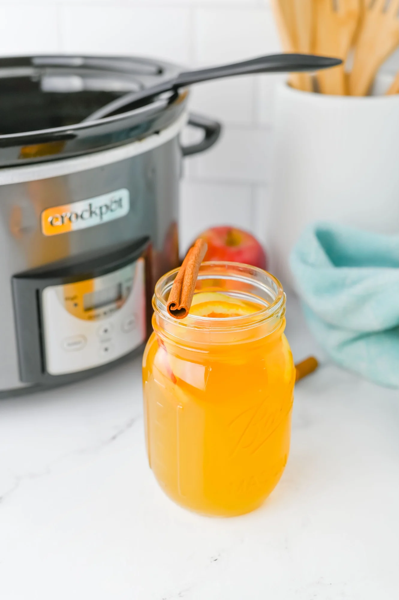 A mason jar filled with orange-yellow liquid and a cinnamon stick is placed in front of a slow cooker on a white kitchen counter.