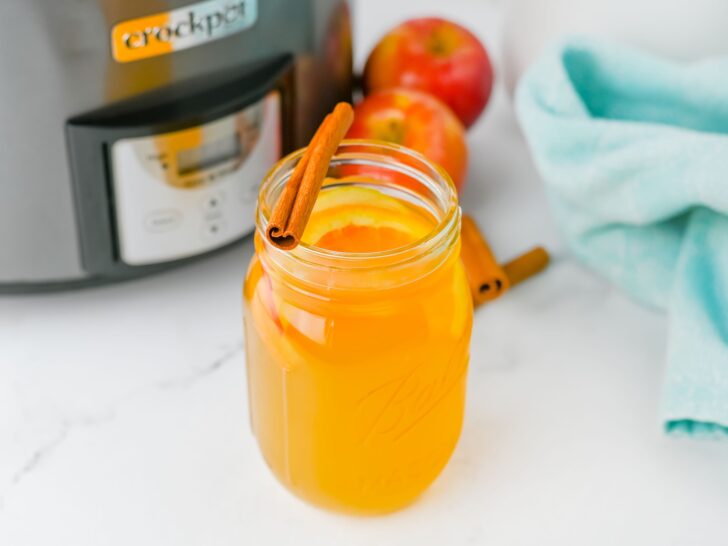 A mason jar filled with crockpot apple cider, garnished with orange slices and a cinnamon stick, sits on a white counter next to fresh apples, a blue towel, and a Crock-Pot.