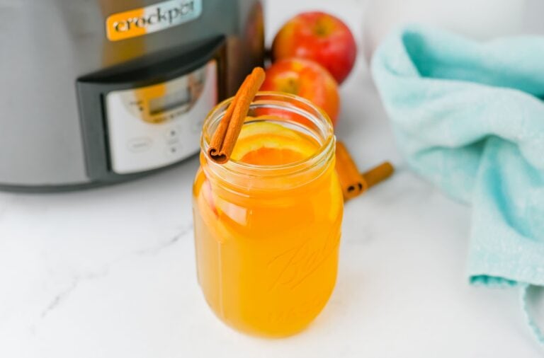 A mason jar filled with crockpot apple cider, garnished with orange slices and a cinnamon stick, sits on a white counter next to fresh apples, a blue towel, and a Crock-Pot.