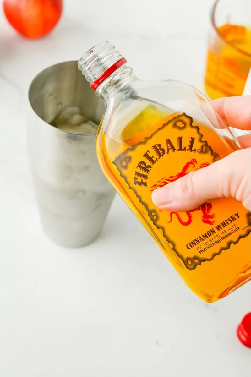 A hand pours Fireball Cinnamon Whisky from a bottle into a metal cocktail shaker filled with ice, with a glass and fruit in the background.