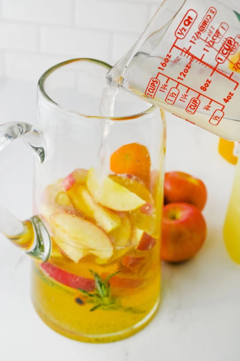 A measuring cup pours liquid into a glass pitcher containing sliced apples, orange, and herbs on a white surface with apples and an orange in the background.