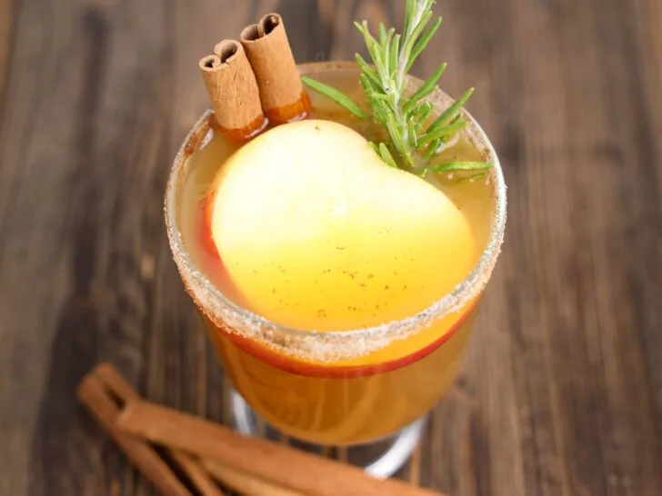 A glass of spiced apple cider garnished with cinnamon sticks, a rosemary sprig, and a slice of apple, set on a wooden surface.