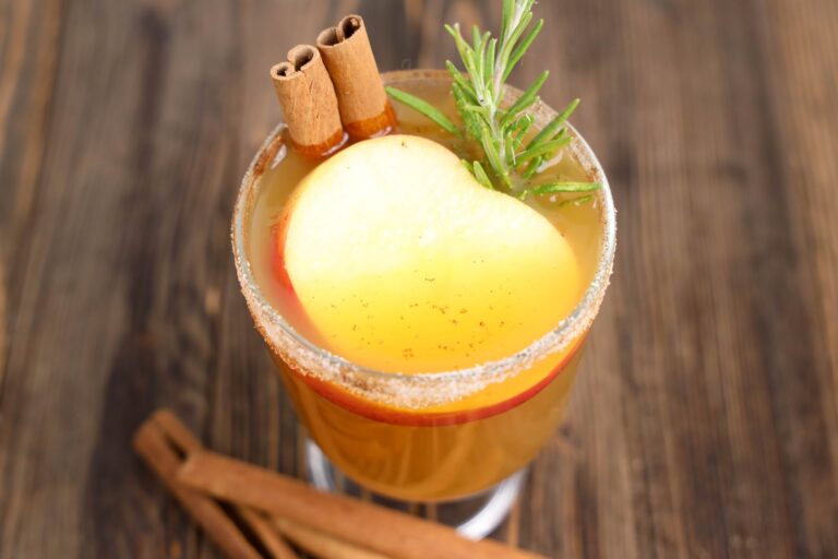 A glass of spiced apple cider garnished with cinnamon sticks, a rosemary sprig, and a slice of apple, set on a wooden surface.
