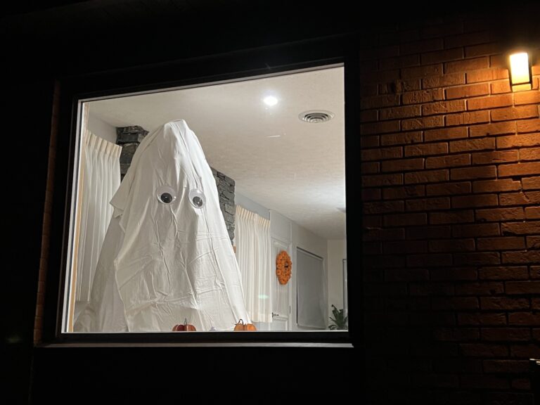 A person wearing a white sheet with large googly eyes stands inside a lit room, viewed through a window at night.