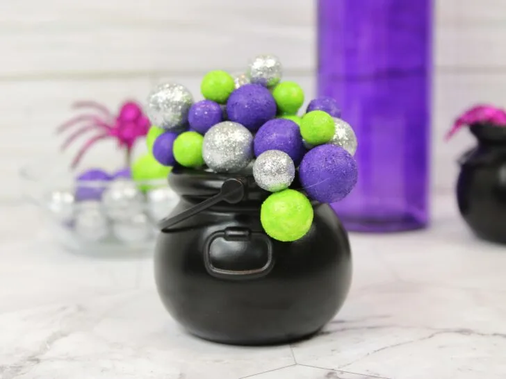 A small black cauldron filled with green, purple, and silver glittery balls sits on a marble surface; decorations and another cauldron are in the background.