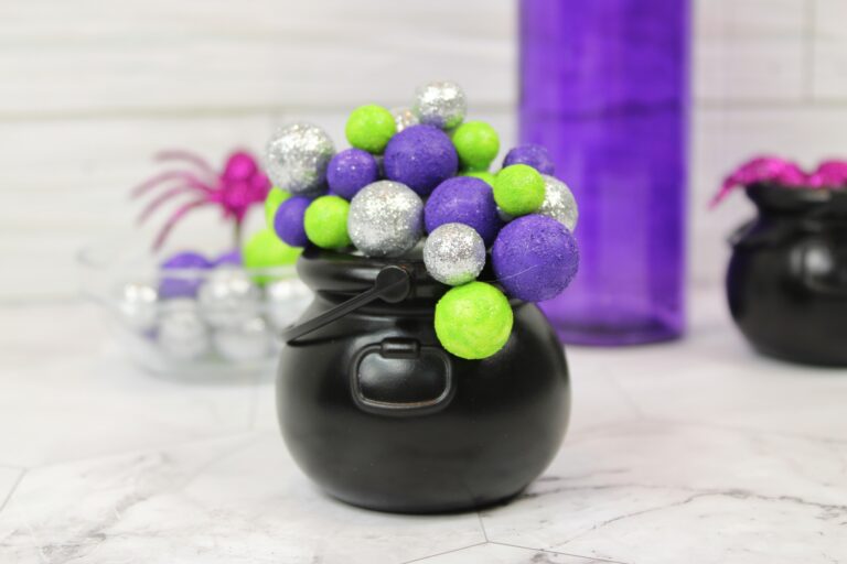 A small black cauldron filled with green, purple, and silver glittery balls sits on a marble surface; decorations and another cauldron are in the background.