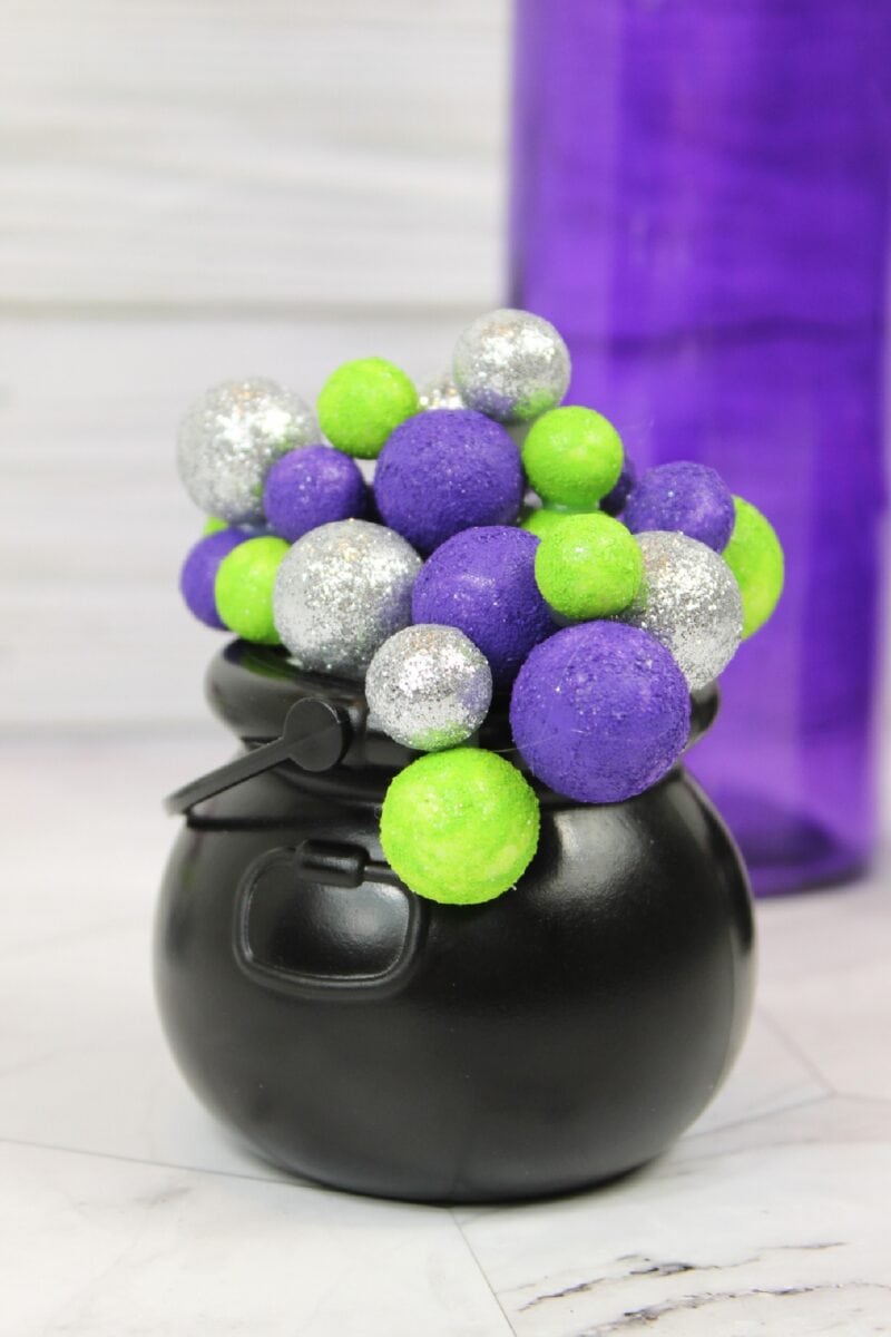 A bubbling cauldron filled with glittery silver, green, and purple foam balls sits on a white surface, with a tall purple object in the background.