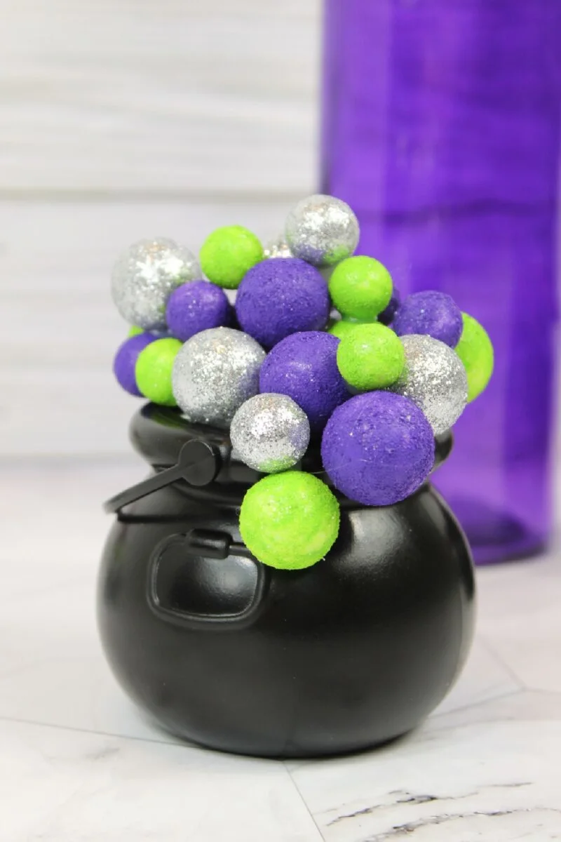 A bubbling cauldron filled with glittery silver, green, and purple foam balls sits on a white surface, with a tall purple object in the background.
