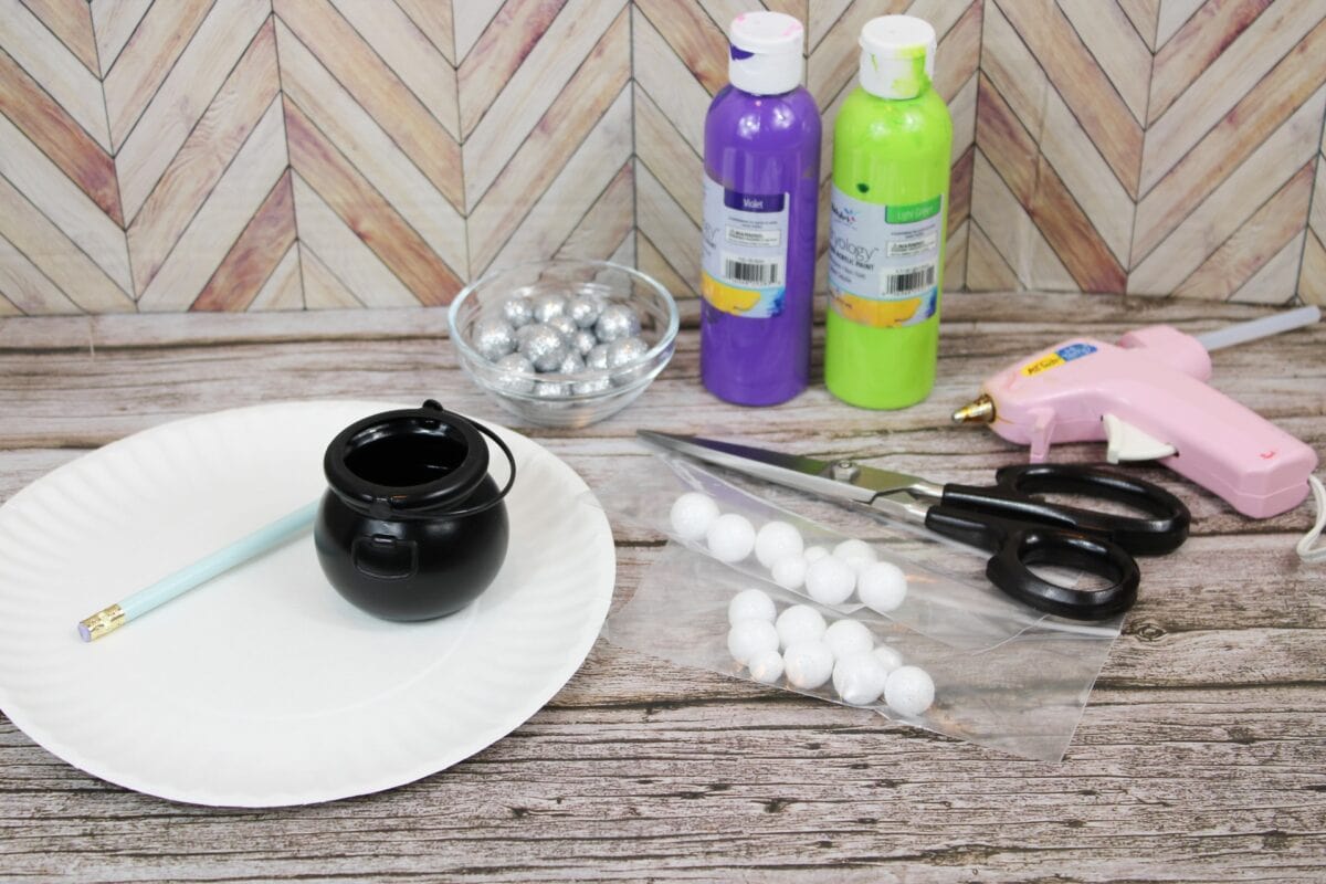 Craft supplies on a wooden table, including a paper plate, pencil, plastic cauldron, scissors, glue gun, paint bottles, silver balls in a bowl, and foam balls in a bag.