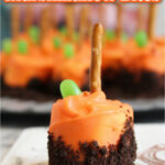 A close-up of a pumpkin patch marshmallow bite with orange frosting, chocolate crumbs, a green candy, and a pretzel stick on top. More pumpkin patch marshmallow bites are blurred in the background.