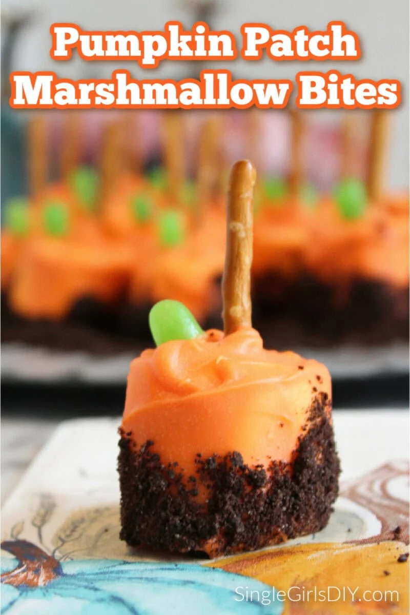 A close-up of a pumpkin patch marshmallow bite with orange frosting, chocolate crumbs, a green candy, and a pretzel stick on top. More pumpkin patch marshmallow bites are blurred in the background.