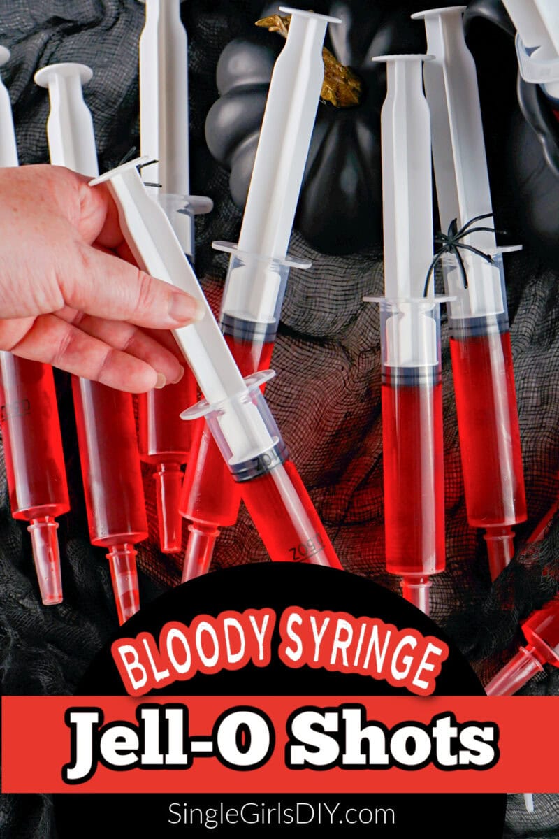 Several plastic syringes filled with red Jell-O are arranged on a dark fabric, with one hand holding a syringe. Text reads, "Syringe Jello Shots Bloody Syringe Jell-O Shots SingleGirlsDIY.com.