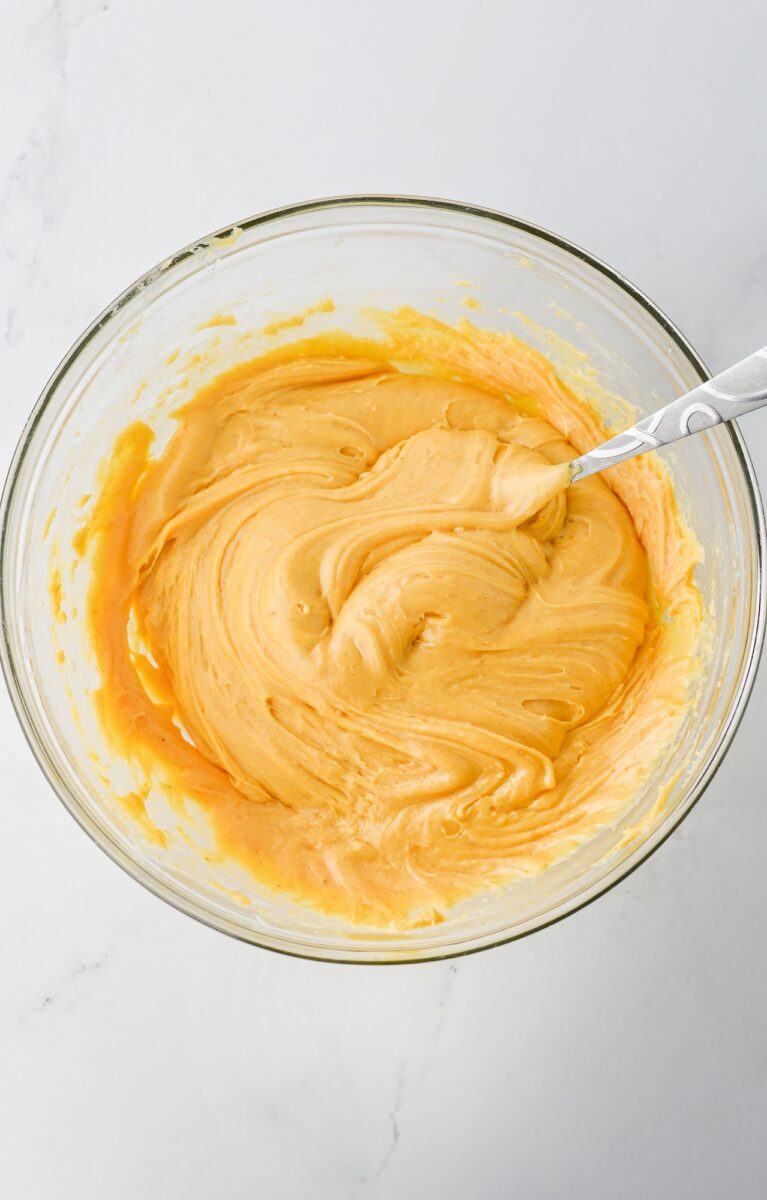 A glass bowl filled with creamy, light orange batter being mixed with a metal spoon on a white surface.