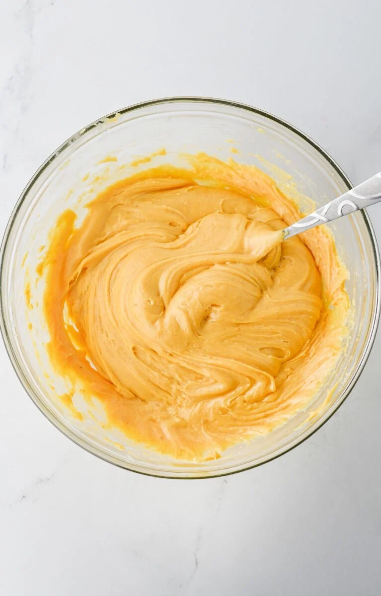 A glass bowl filled with creamy, light orange batter being mixed with a metal spoon on a white surface.