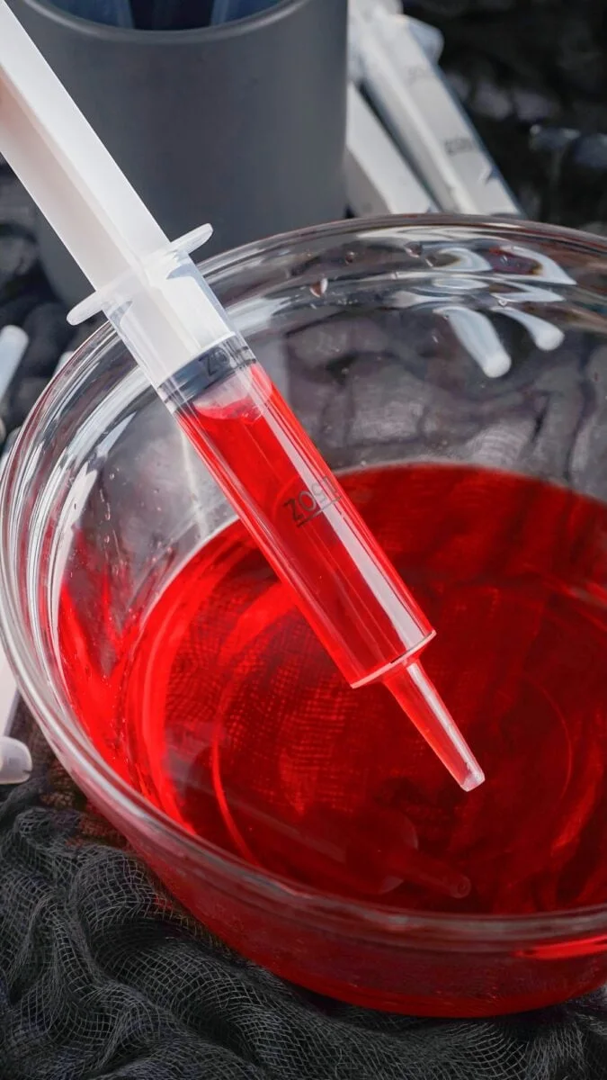 A clear syringe filled with red liquid rests on the rim of a glass bowl containing more red syrup, suggesting fun syringe jello shots ready to be enjoyed.