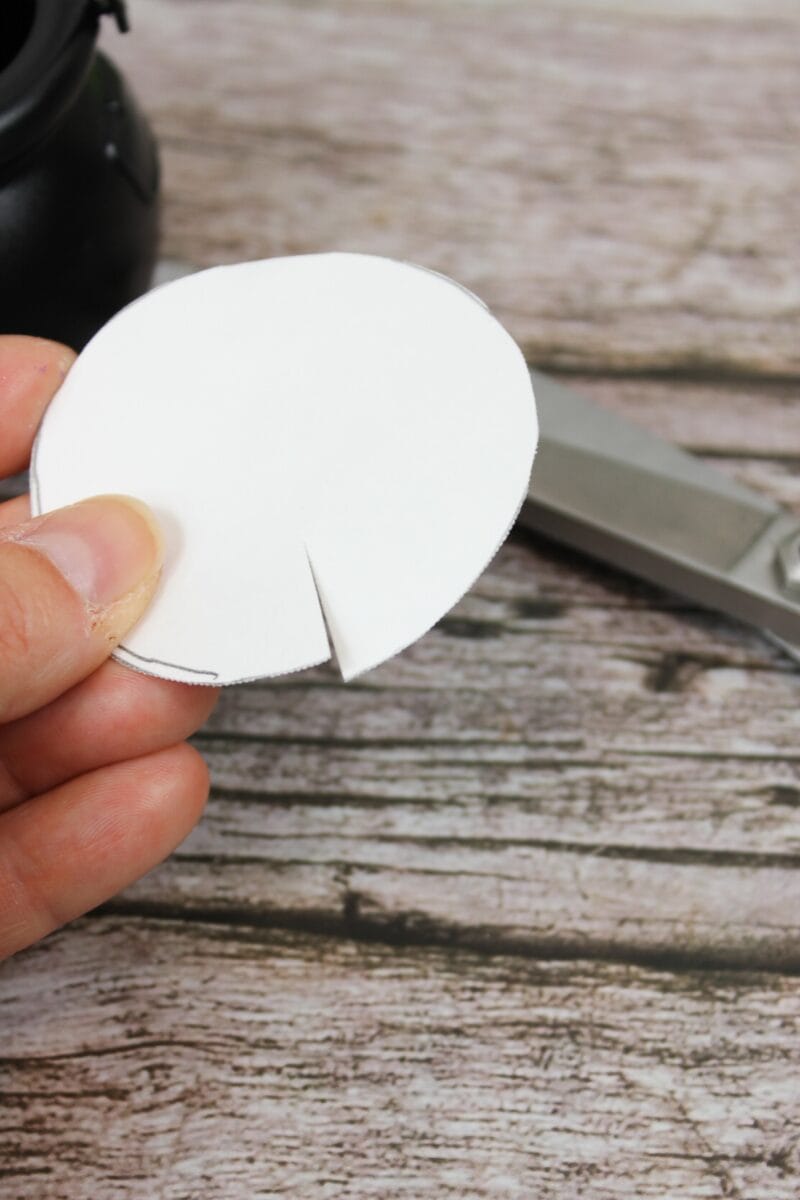 A hand holds a white circular piece of paper with a single straight cut, next to a pair of scissors on a wooden surface.