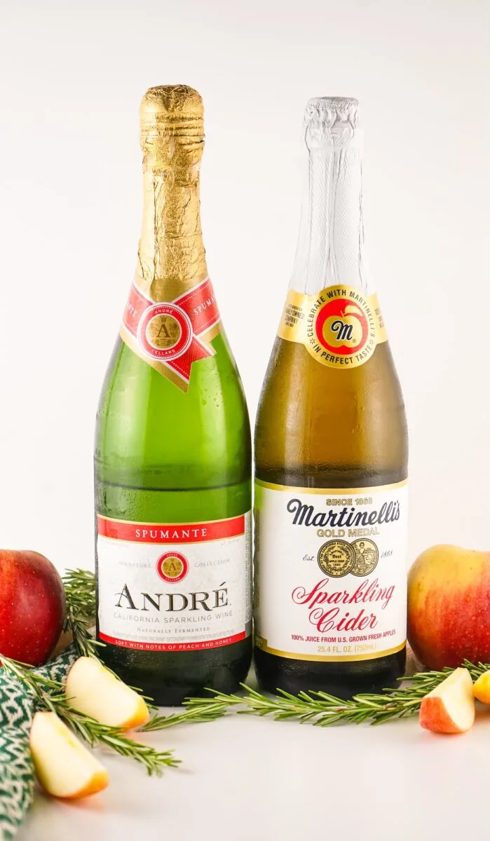 Two bottles, one labeled "André Spumante California Sparkling Wine" and the other "Martinelli's Gold Medal Sparkling Cider," are placed side by side with apples and greenery around them.