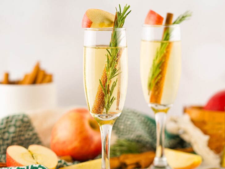 Two champagne glasses filled with a light beverage, each garnished with a sprig of rosemary, a cinnamon stick, and an apple slice. Apples and cinnamon sticks are in the background.