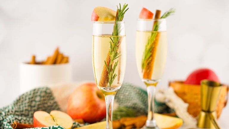 Two champagne glasses filled with a light beverage, each garnished with a sprig of rosemary, a cinnamon stick, and an apple slice. Apples and cinnamon sticks are in the background.