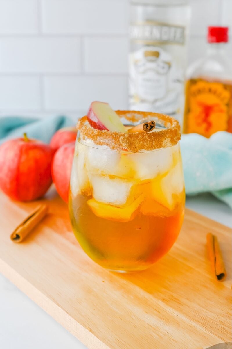 A glass of iced apple pie cocktail with a brown sugar rim, garnished with apple slices and a cinnamon stick, sits on a wooden board surrounded by apples, cinnamon sticks, and bottles in the background.