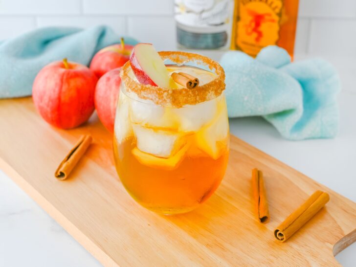 A glass of iced apple pie cocktail with a cinnamon stick and apple slice garnish, surrounded by apples and cinnamon sticks on a wooden tray.