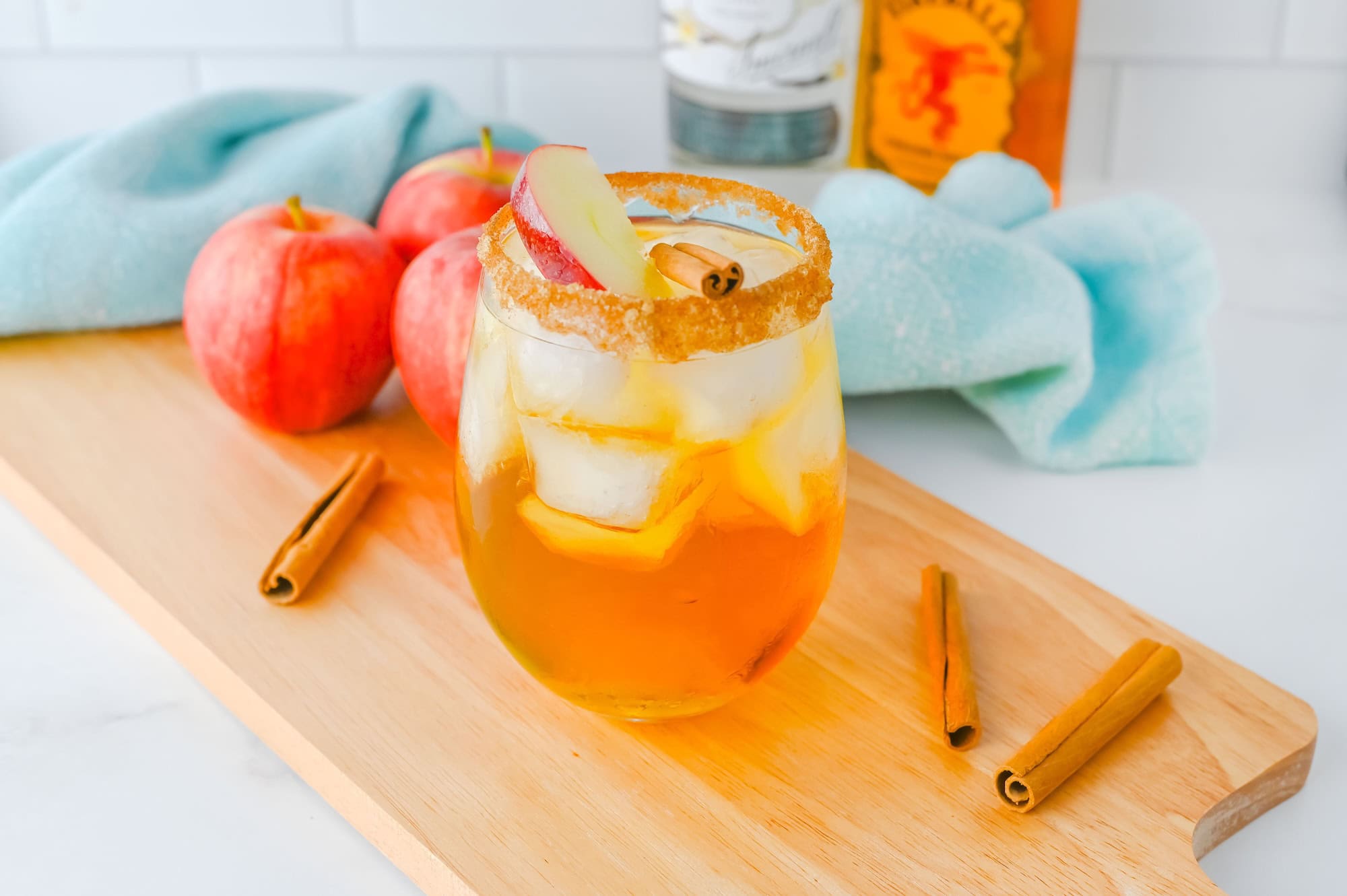 A glass of iced apple pie cocktail with a cinnamon stick and apple slice garnish, surrounded by apples and cinnamon sticks on a wooden tray.