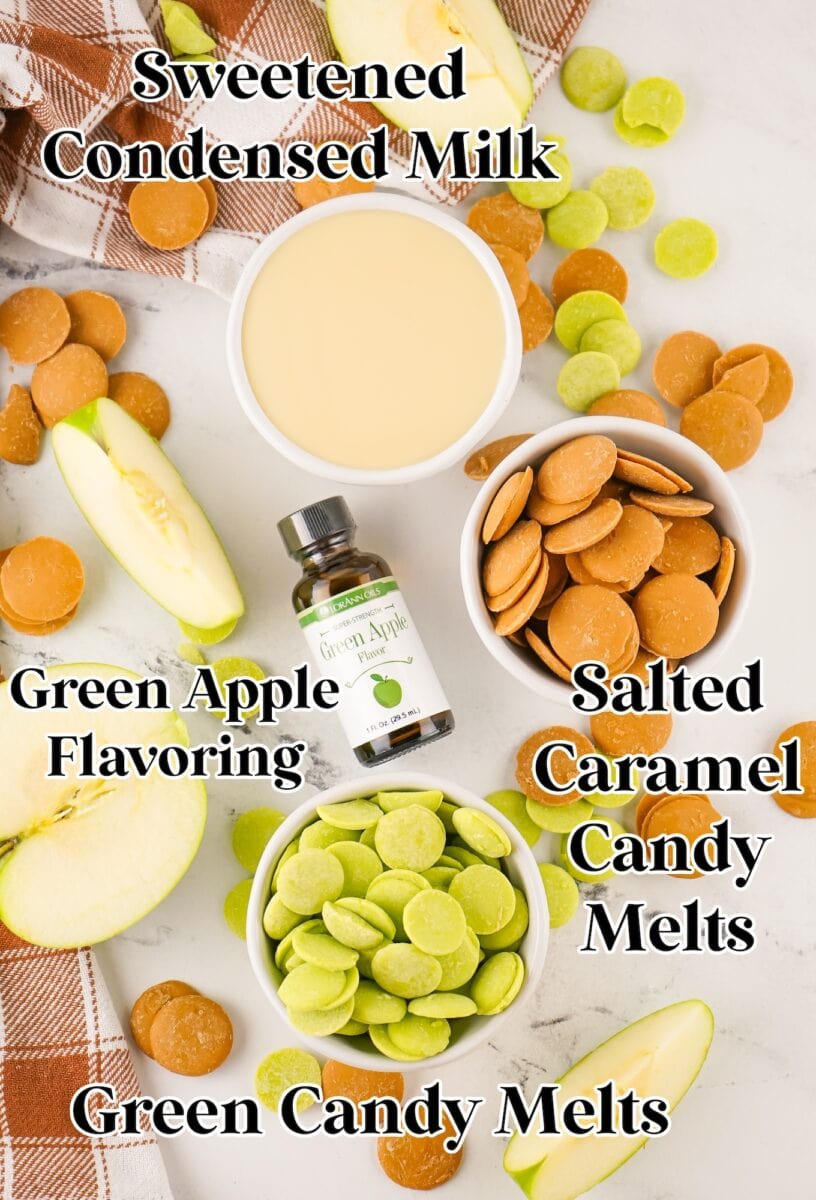 Flat lay of ingredients for caramel apple fudge, including sweetened condensed milk, green apple flavoring, green and salted caramel candy melts, apple slices, and a plaid cloth on a marble surface.